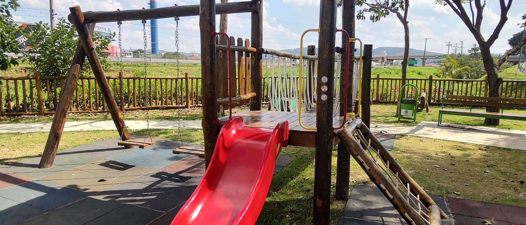 Torre de playground em toras de eucalipto com telhado, escorregador vermelho, balanço e piso de placas de borracha em praça de condomínio